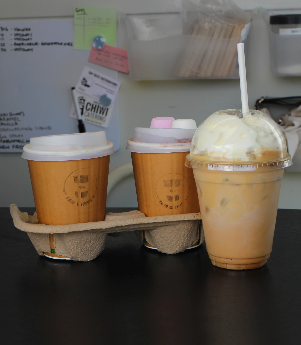 A cup of coffee, a cup of hot chocolate with two marshmallows on it, and an iced coffee on a bench.